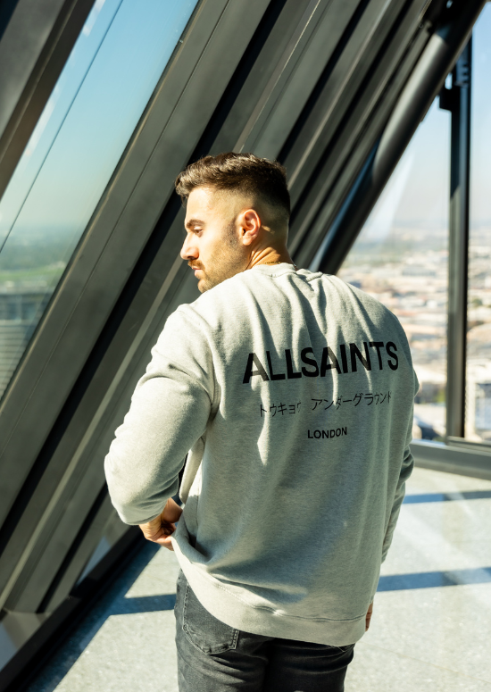 A person wearing a gray AllSaints sweatshirt stands by large windows, overlooking a cityscape, as if stepping into a scene from Enrico Incarnati Productions.
